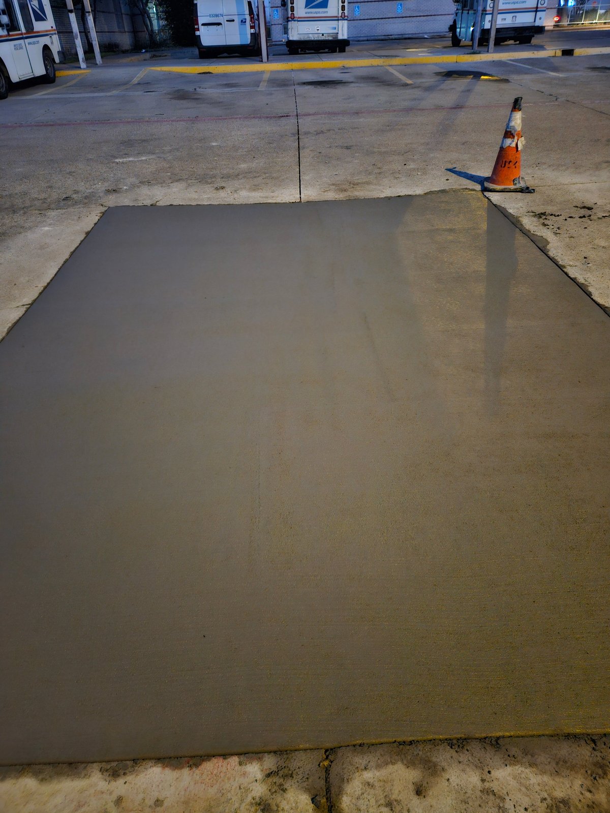 Freshly poured concrete slab on a construction site with orange traffic cone and parked vehicles in background