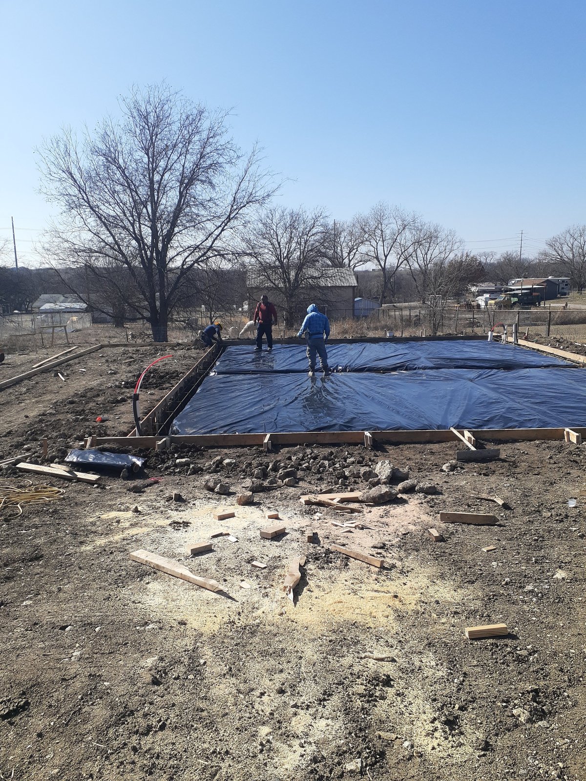 Crew installing a blue vapor barrier over a foundation in a rural property with bare trees and dirt ground visible