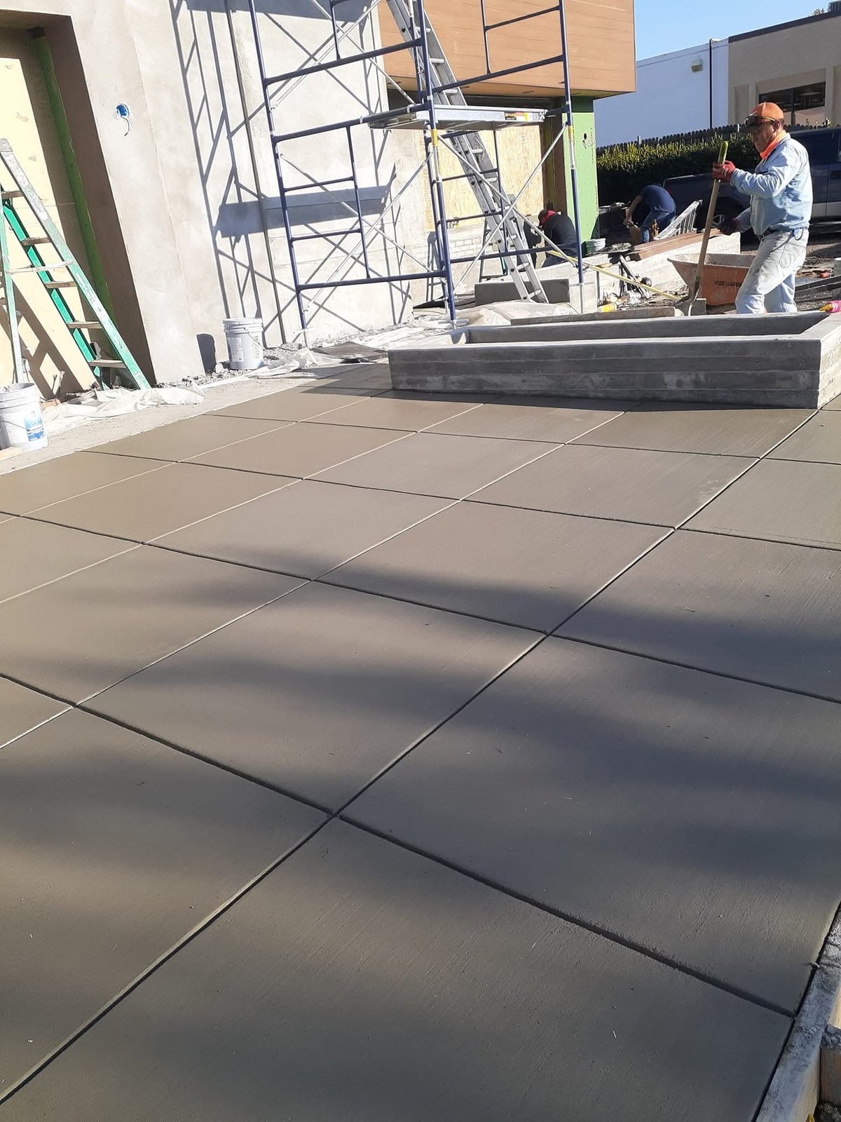 Concrete patio under construction with workers and metal scaffolding visible in background on sunny day