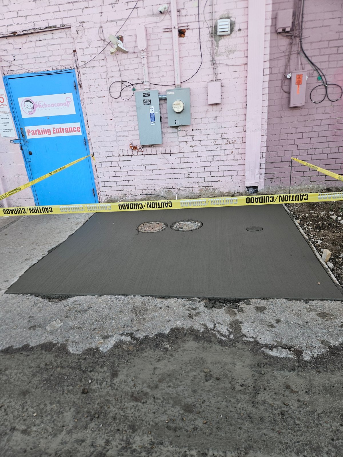 Freshly poured concrete patch at a parking entrance with caution tape, against a brick wall with electrical equipment