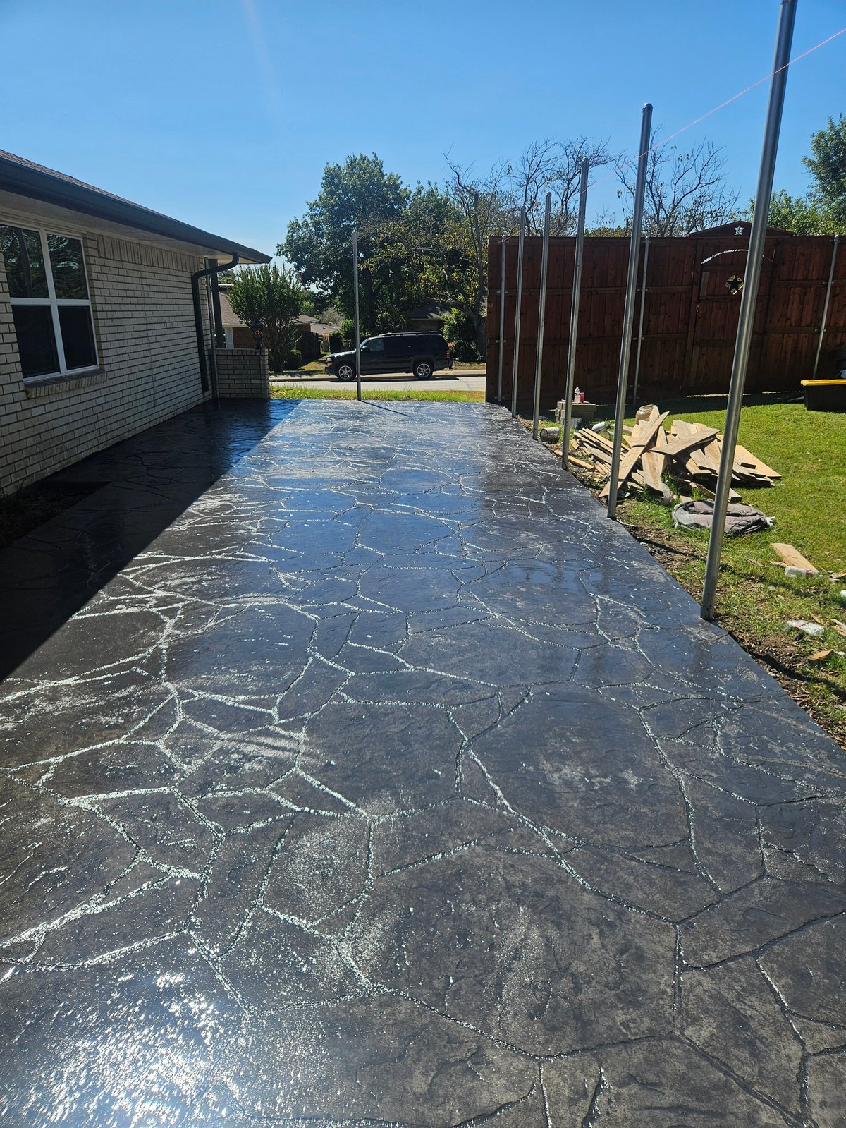 Stamped concrete patio extending from brick house with red wooden fence and metal support posts in sunny backyard