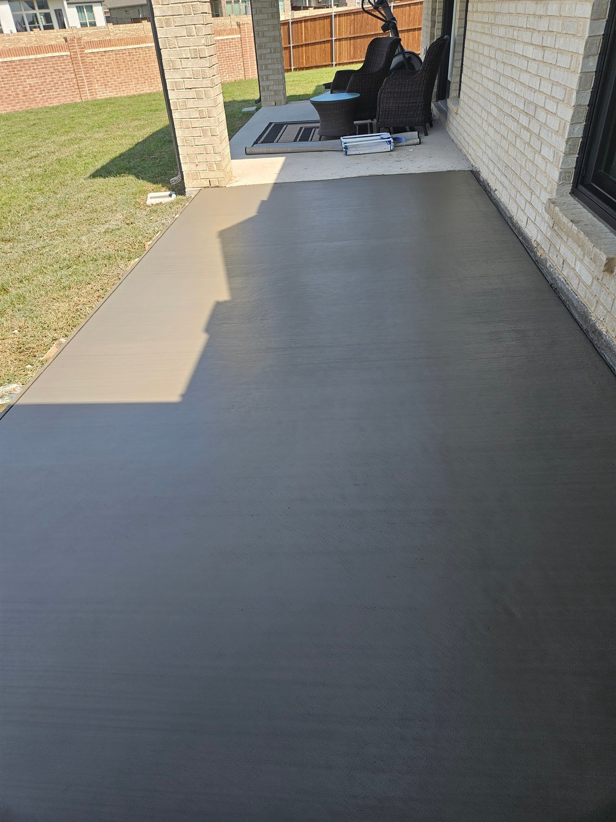 Newly poured dark concrete patio extending from brick house with lawn and fence visible in background