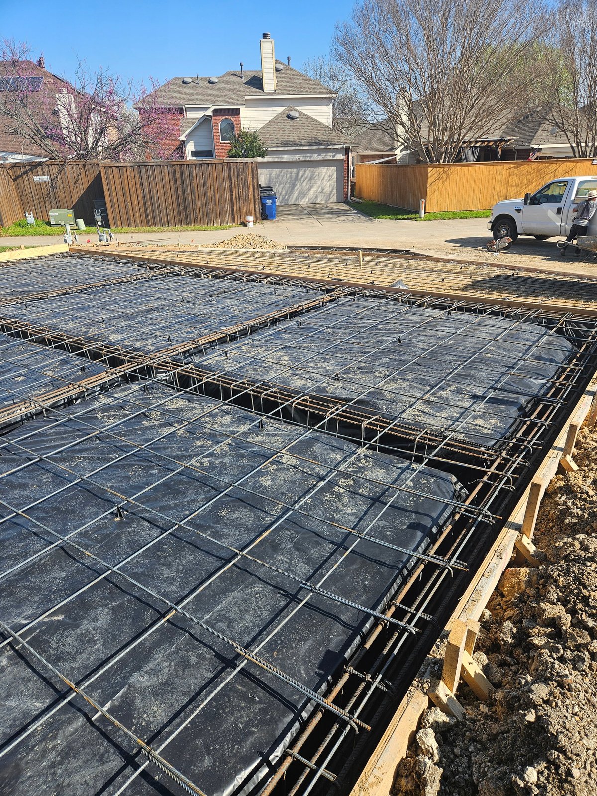 Construction site with reinforced concrete foundation and rebar framework laid out in a residential backyard with a house visible in the background