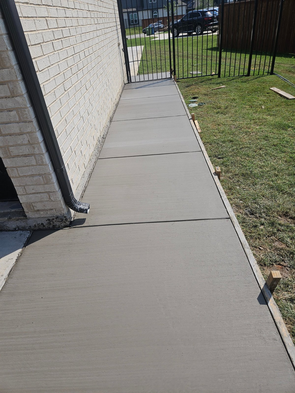 Concrete pathway along brick house exterior leading to black metal gate, with green lawn and parked car visible