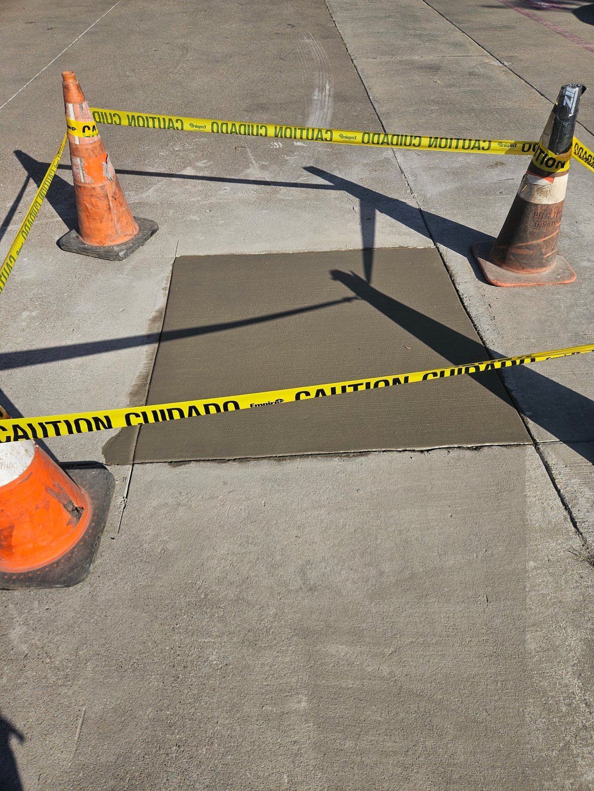 Construction area with yellow caution tape cordoning off a rectangular open hole in concrete pavement, surrounded by traffic cones
