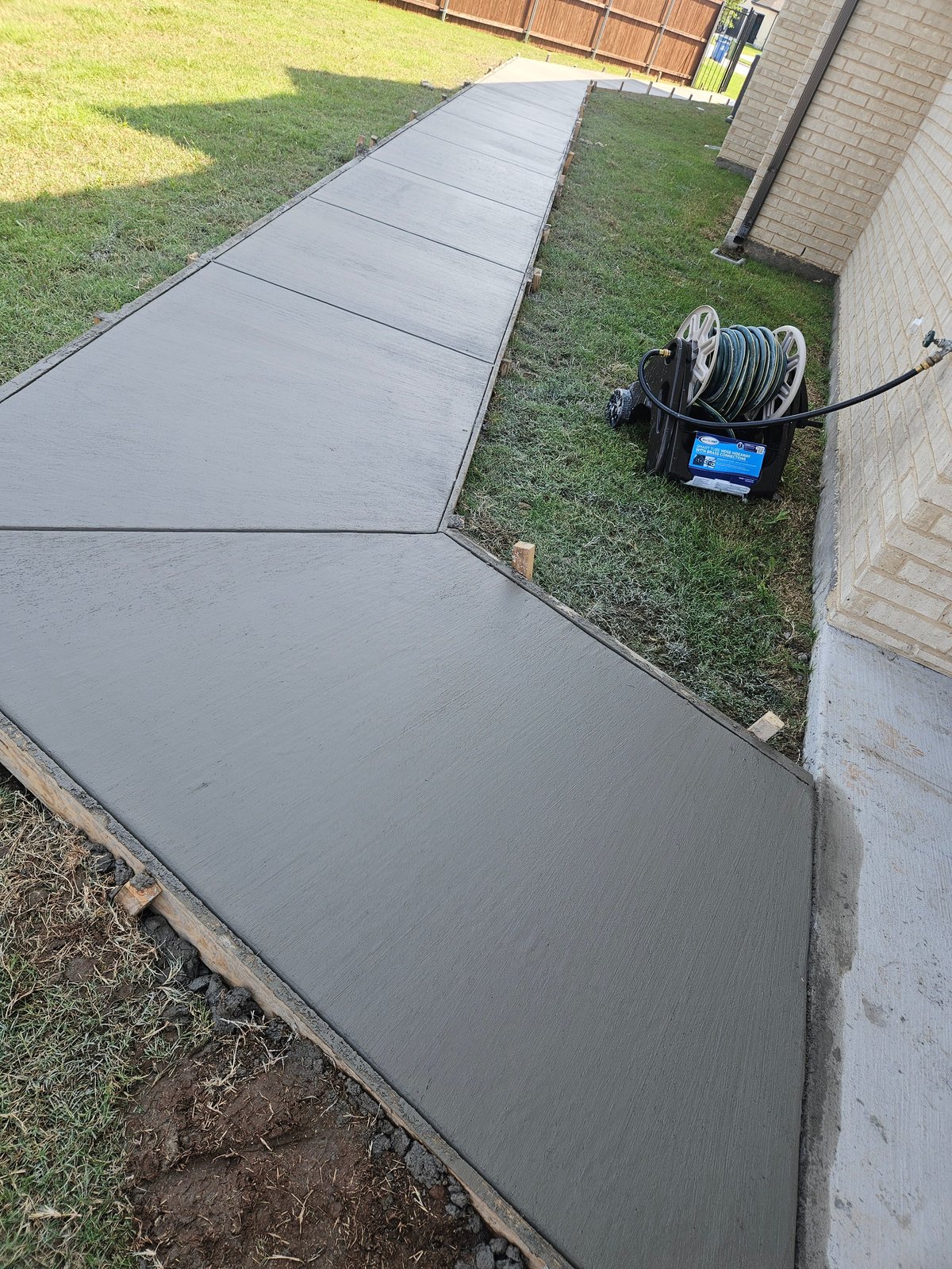 Concrete pathway being freshly poured alongside a brick house with green lawn and garden hose reel visible