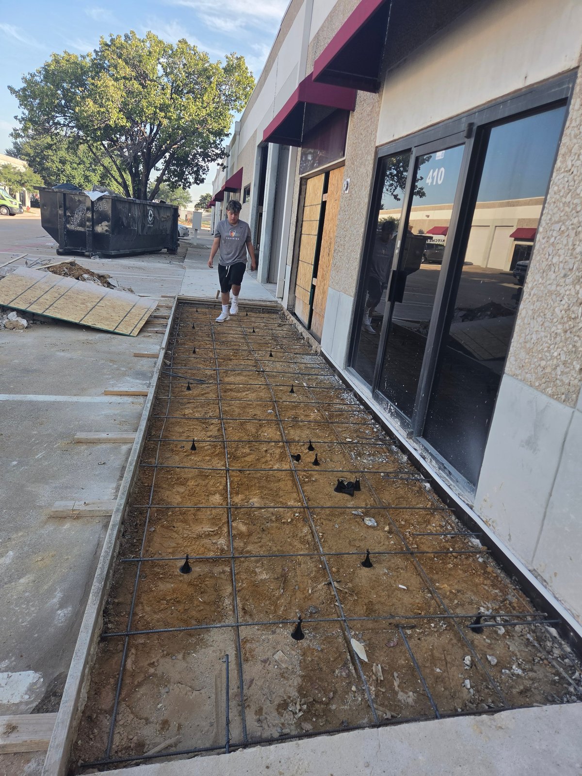 Construction site with exposed soil and grid layout in front of modern building with black windows and person walking in background
