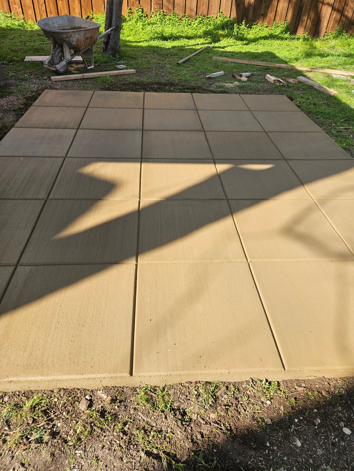 Concrete patio tiles in a backyard with tree shadow, wooden fence, and lawn in background