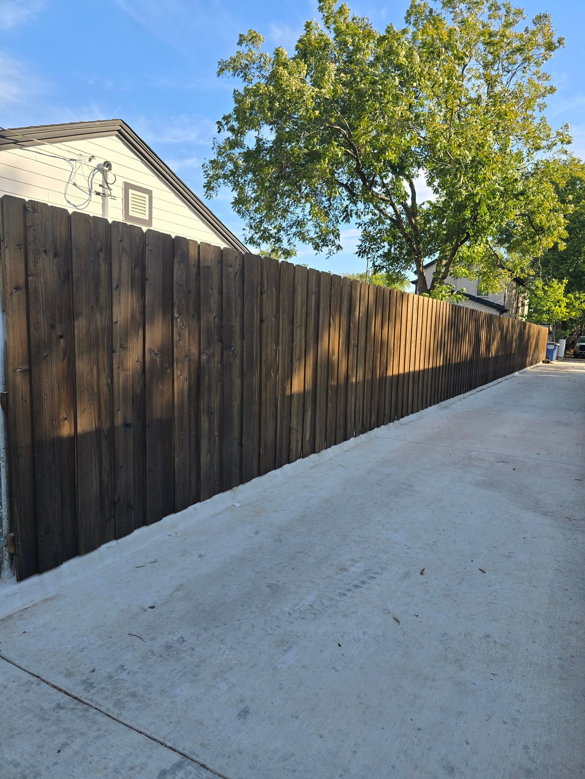 Dark wooden privacy fence lining a concrete driveway with a house and large green tree visible in the background under blue sky