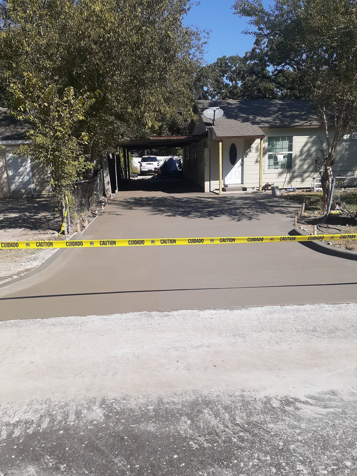 Single-story residential house with yellow caution tape across the driveway entrance, white vehicle visible in garage, trees surrounding property on clear day