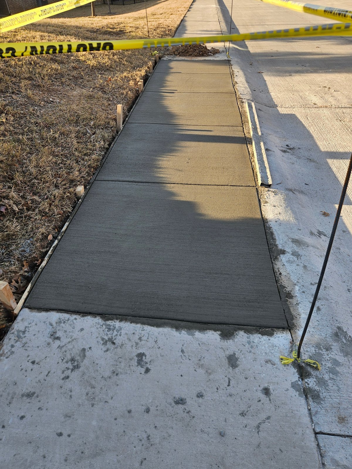 Newly poured concrete walkway with caution tape, connecting two sections of existing sidewalk next to a gravel area