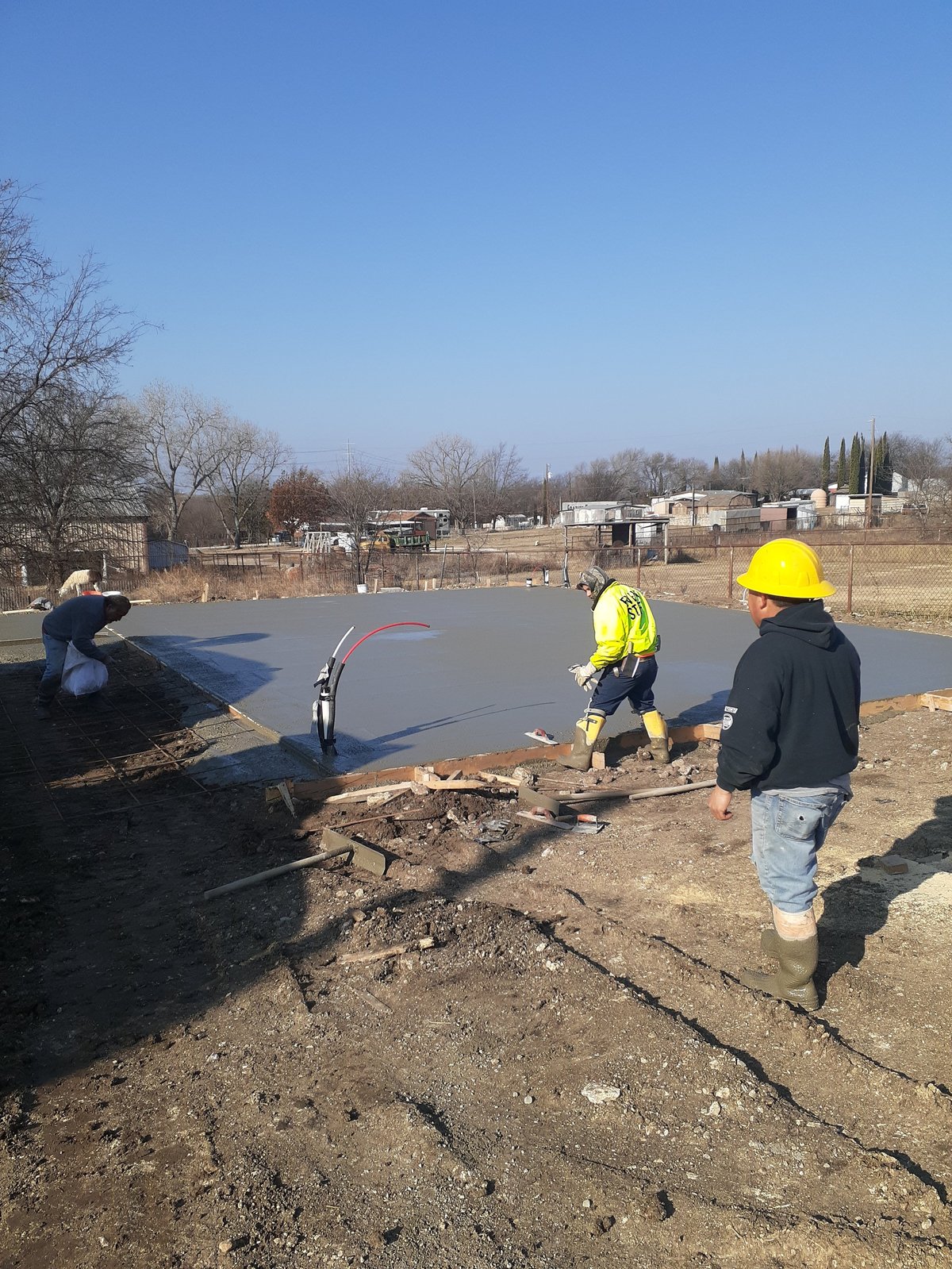 Construction workers installing dark asphalt or roofing material on a flat surface in a rural area with bare trees and buildings in the background