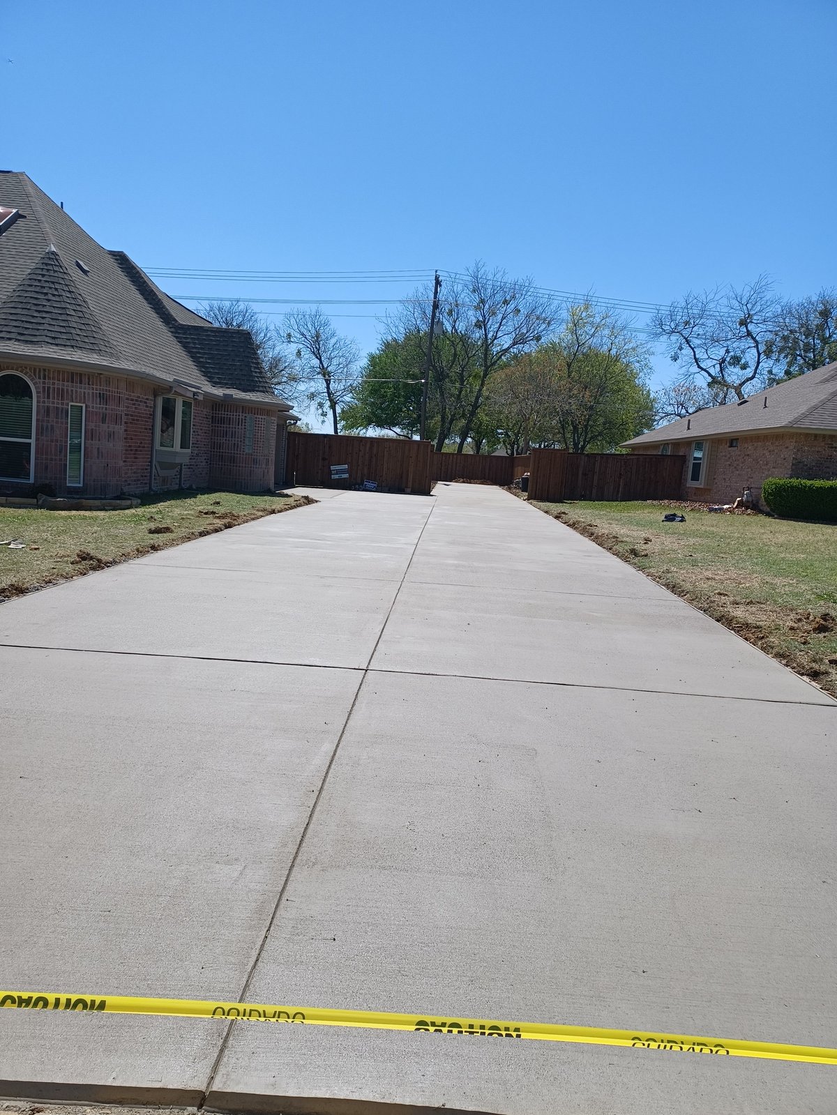 Suburban residential driveway with brick houses, bare and green trees, clear sky, and yellow caution tape across the foreground