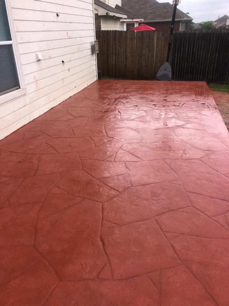 Red stamped concrete patio in backyard with white house siding, dark wooden fence, and red umbrella visible