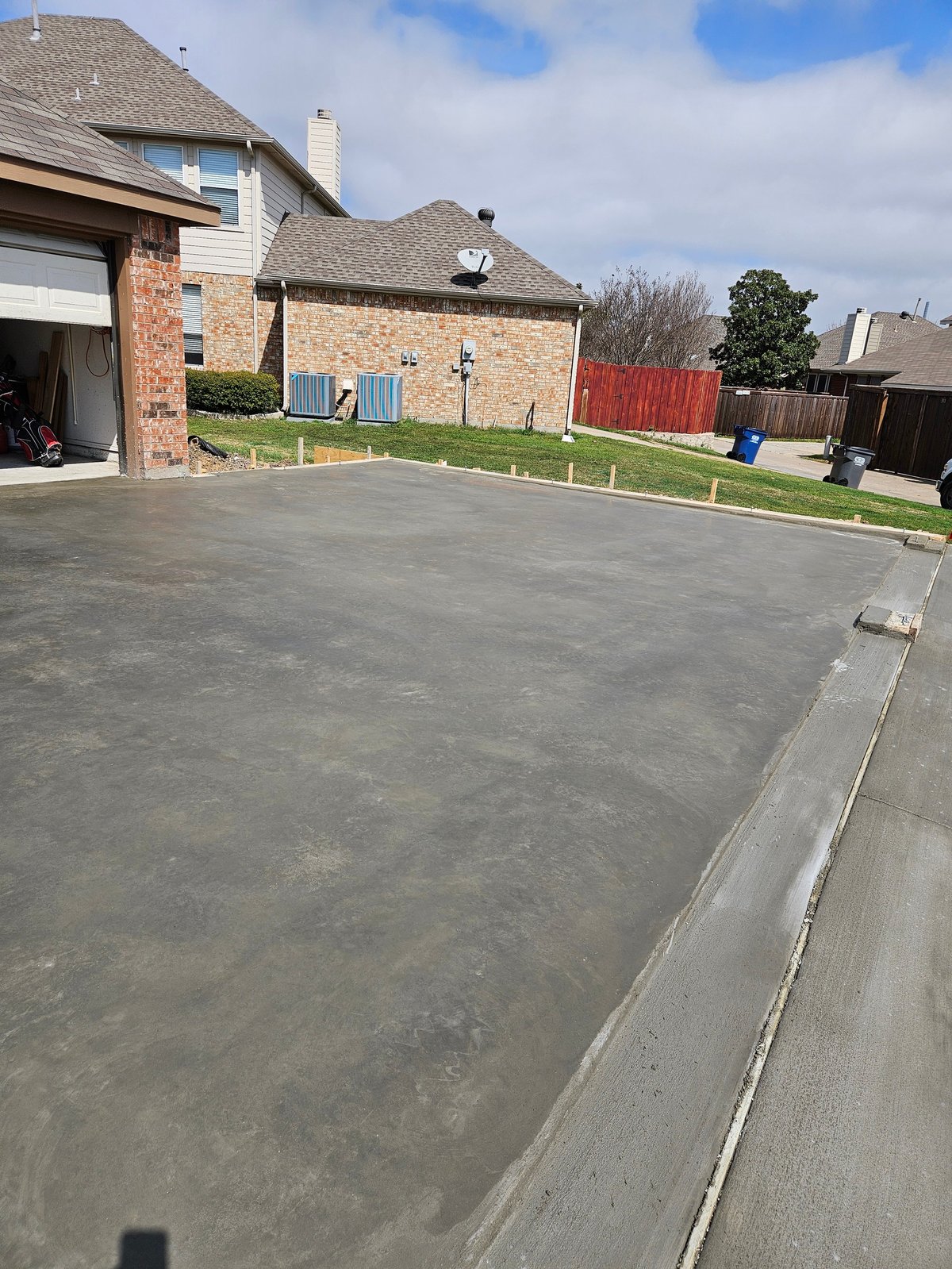 Newly poured concrete driveway in front of two-story brick house with garage, green lawn, and residential fencing