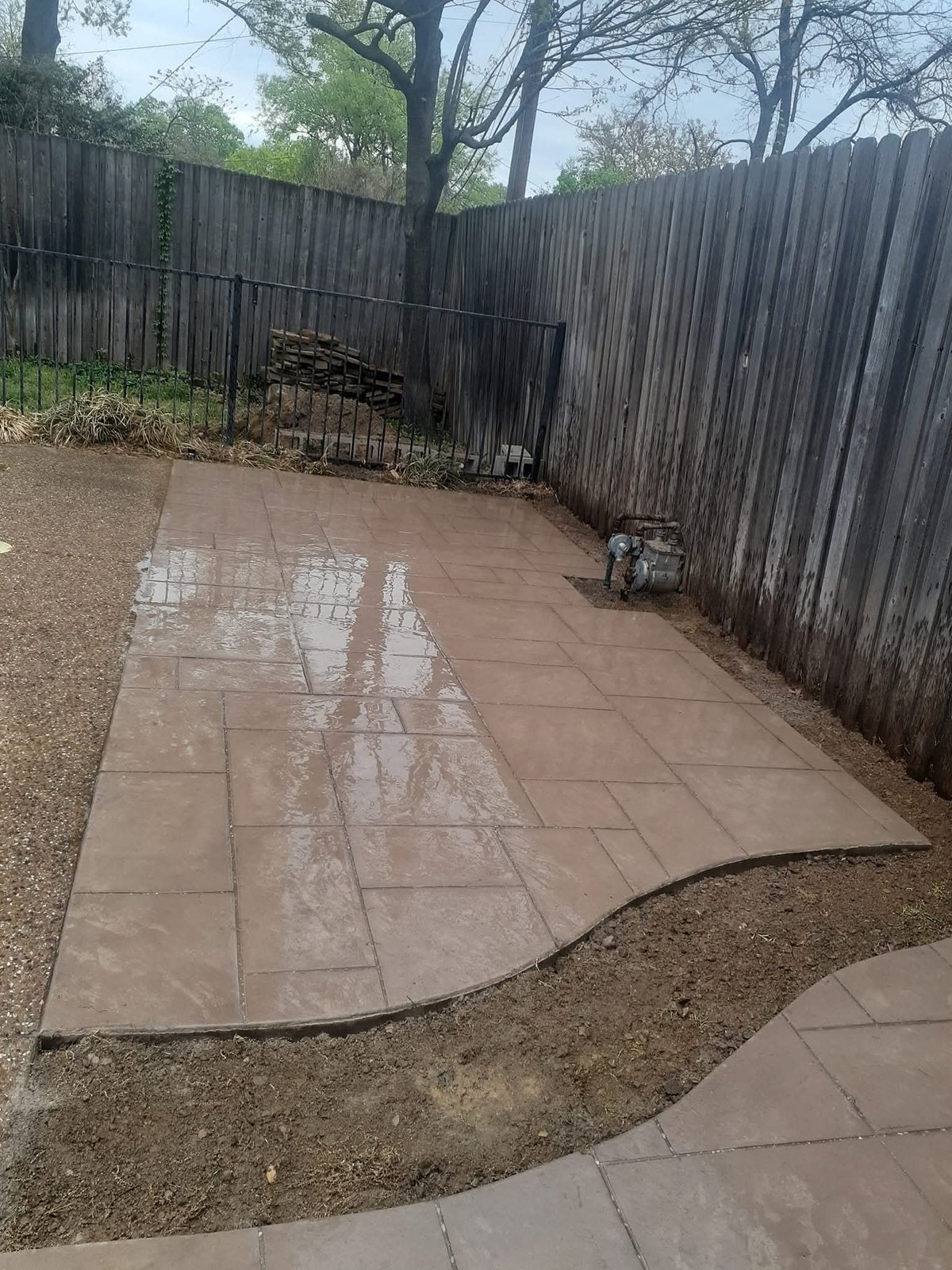 Wet patio with puddles reflecting sky, bordered by weathered wooden fence and bare trees in residential backyard