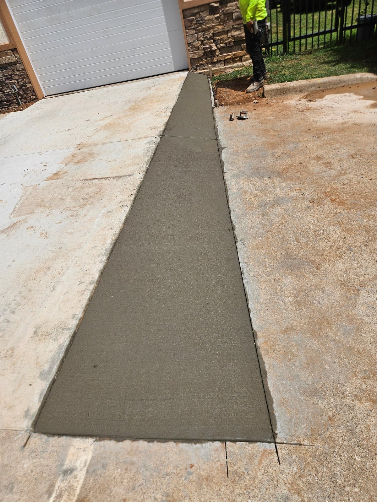 Freshly poured concrete patch connecting a driveway to a dirt area beside a garage, with landscaping visible in background