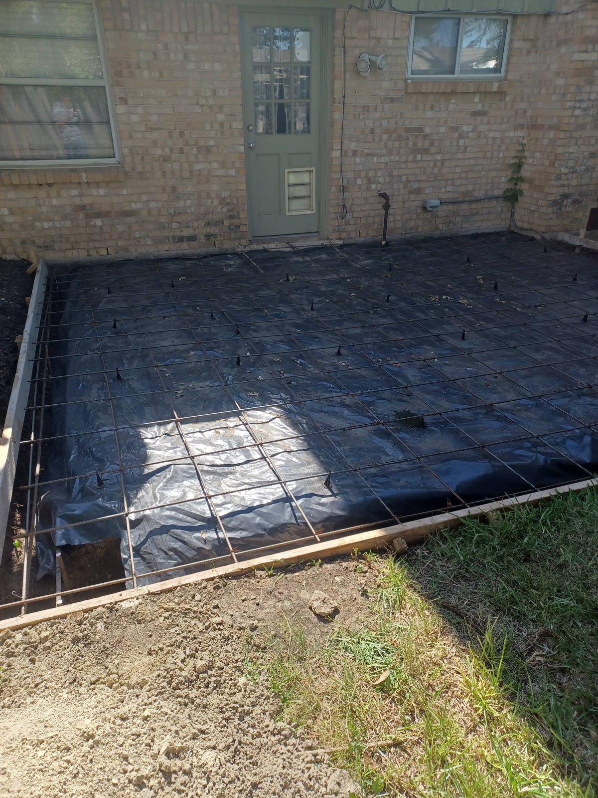 Outdoor patio area with rebar grid and black plastic sheeting secured to ground, brick building and door in background