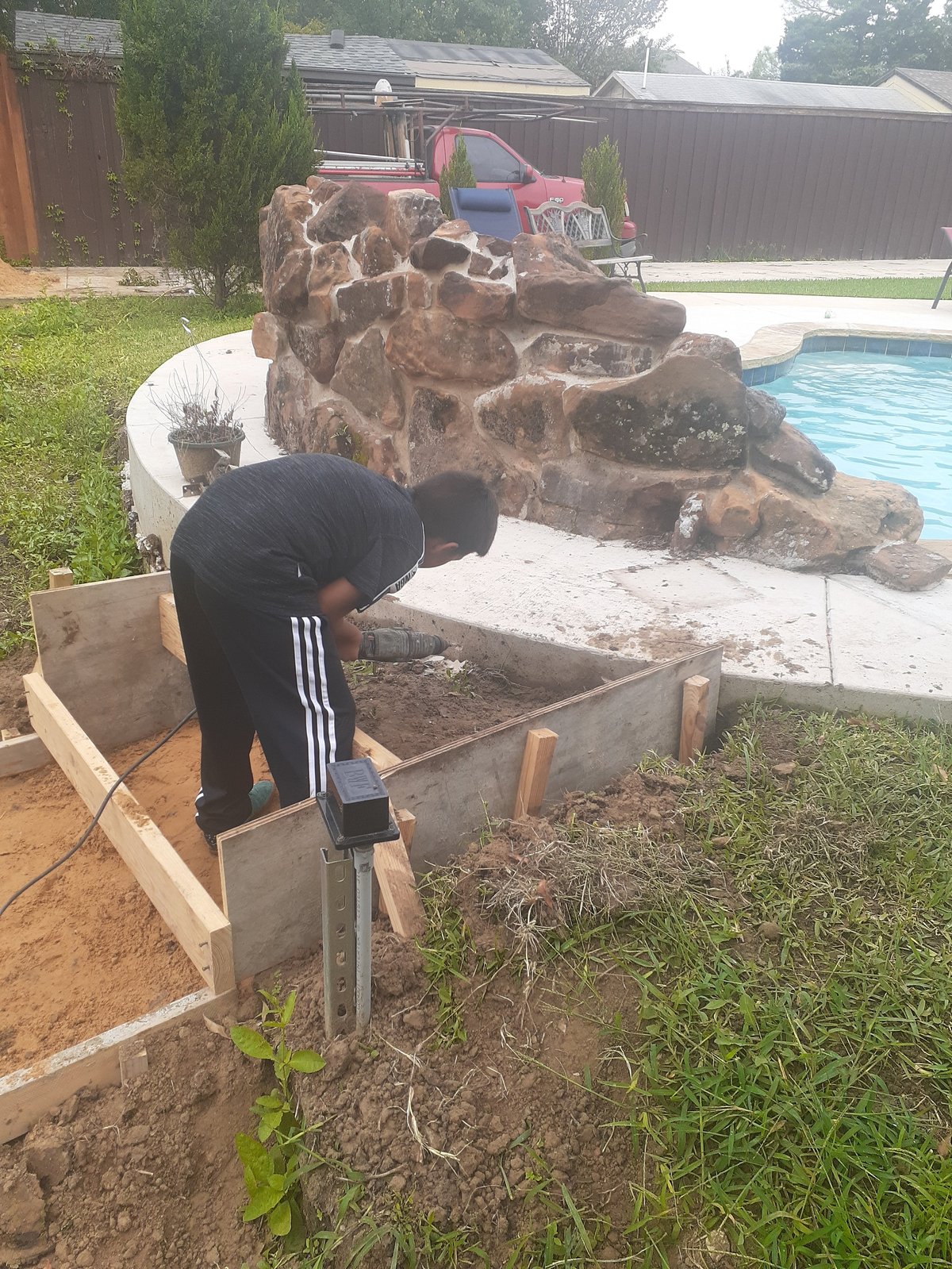 Person digging in a raised garden bed with a shovel, with a large rock formation and swimming pool visible in the background