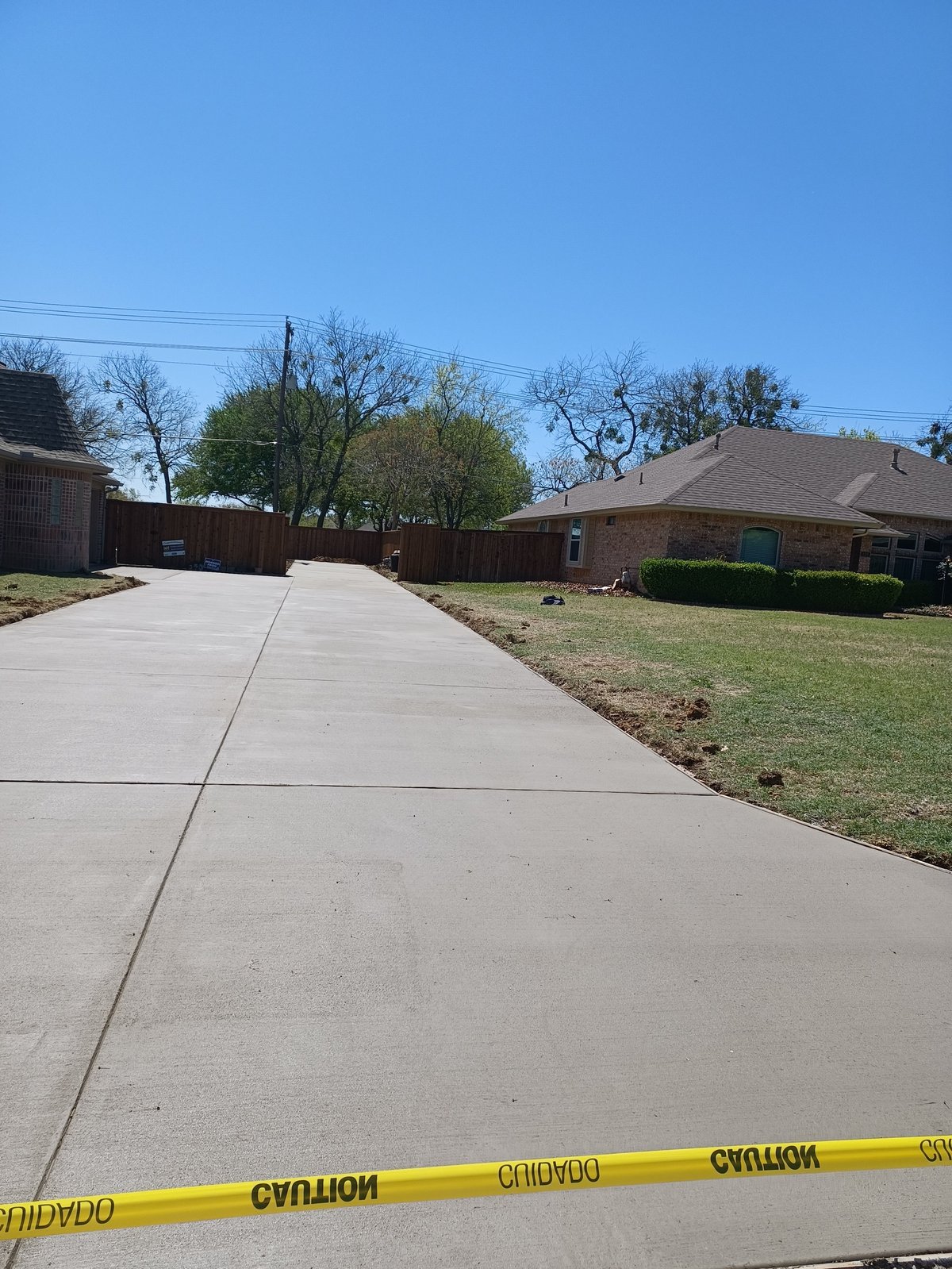Long residential concrete driveway with caution tape across the foreground, single-story brick house and lawn in the background
