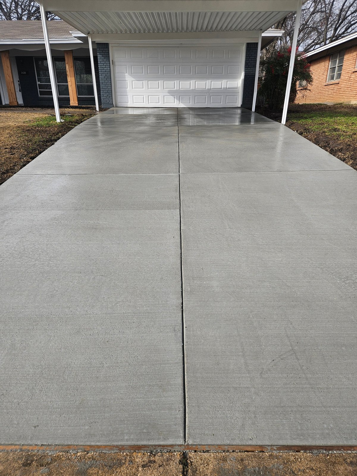 Empty residential driveway with white garage door, expansion joint running down the center, flanked by brick house and carport
