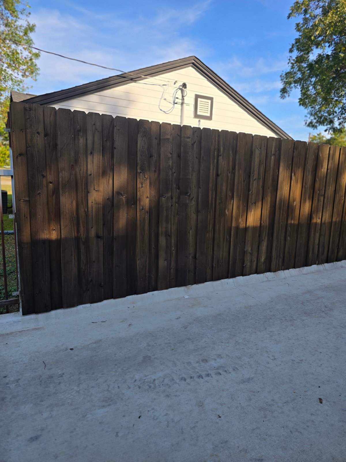 Dark wooden privacy fence with cream-colored house behind it, concrete driveway in foreground, blue sky with trees visible