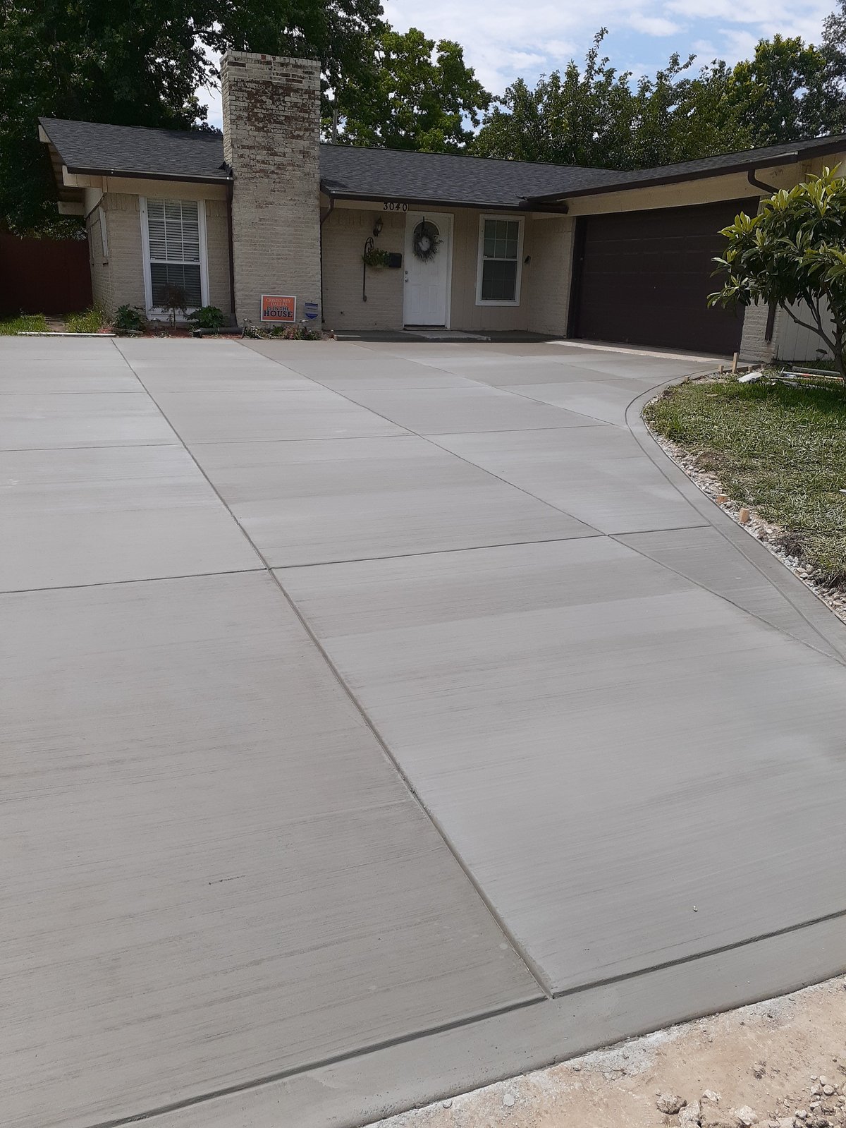 Single-story residential home with cream brick facade, dark roof, and new concrete driveway under clear sky